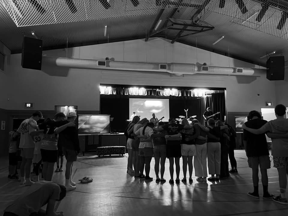 Black and white photo of a crowd gathered arm in arm during a song in a hall